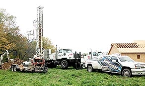 Photo of white rotary drilling rig, water truck, skid loader and dually truck at a new home construction site 
